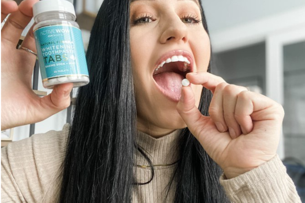 Young woman holding Active Wow Toothpaste Tabs in one hand and holding one tab in the other hand near her mouth